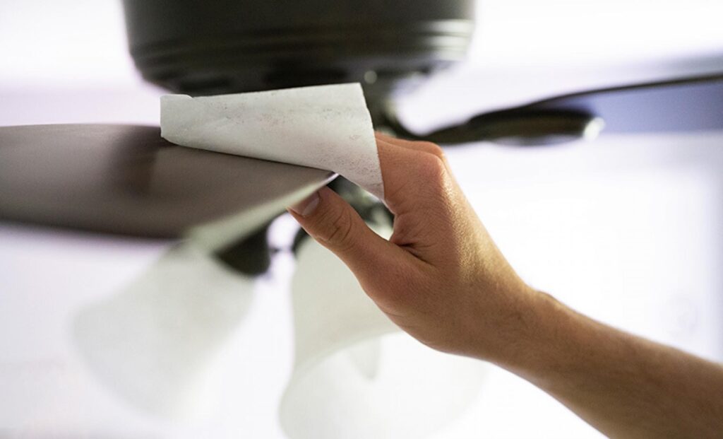 Close de uma pessoa usando uma fronha branca para limpar a pá de um ventilador de teto, mostrando como as pessoas que usam a fronha para limpar evitam a sujeira.